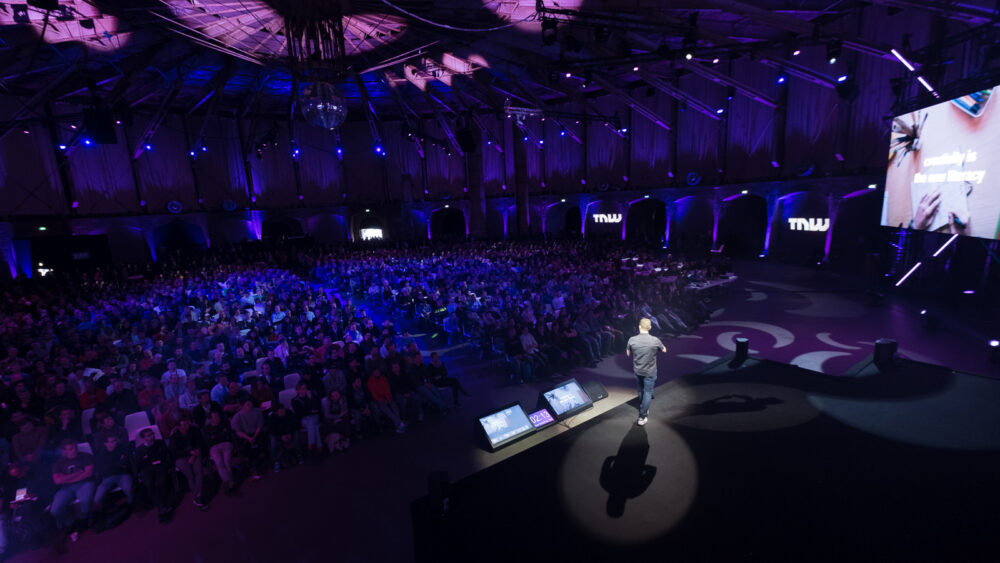 Chase Jarvis on stage at The Next Web Conference, addressing a large audience in a dimly lit auditorium with purple lighting. The audience is seated, attentively listening to the presentation. The stage is equipped with large screens displaying slides, and the venue has a modern, high-tech atmosphere.