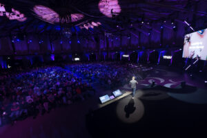 Chase Jarvis on stage at The Next Web Conference, addressing a large audience in a dimly lit auditorium with purple lighting. The audience is seated, attentively listening to the presentation. The stage is equipped with large screens displaying slides, and the venue has a modern, high-tech atmosphere.