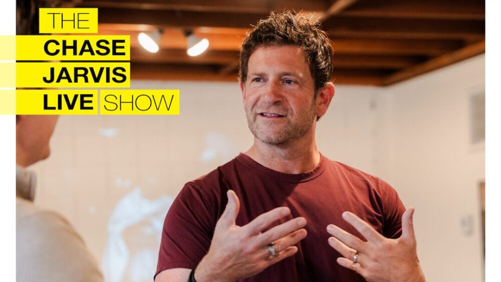 Chase Jarvis in mid-conversation, gesturing with his hands, wearing a casual maroon t-shirt. The background appears to be an indoor space with wooden beams on the ceiling and soft lighting. On the left side, a bold yellow and black graphic reads "THE CHASE JARVIS LIVE SHOW," with each word stacked in a column. The scene conveys an energetic and engaging atmosphere, with Chase passionately explaining something to the person in front of him.